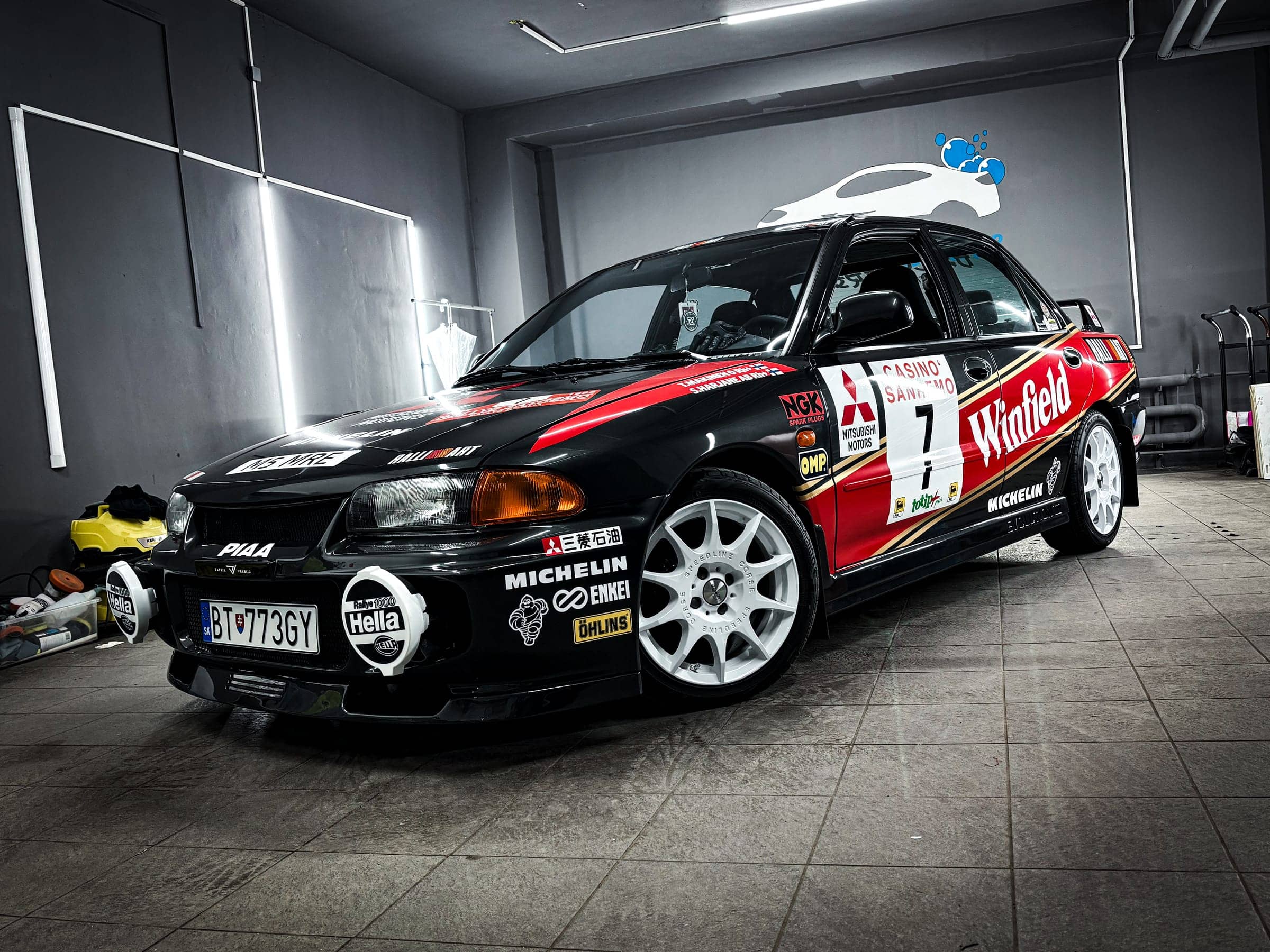 Mitsubishi Lancer Evolution s rally polepom Winfield, štartovým číslom 7 a logami sponzorov v tmavom štúdiu